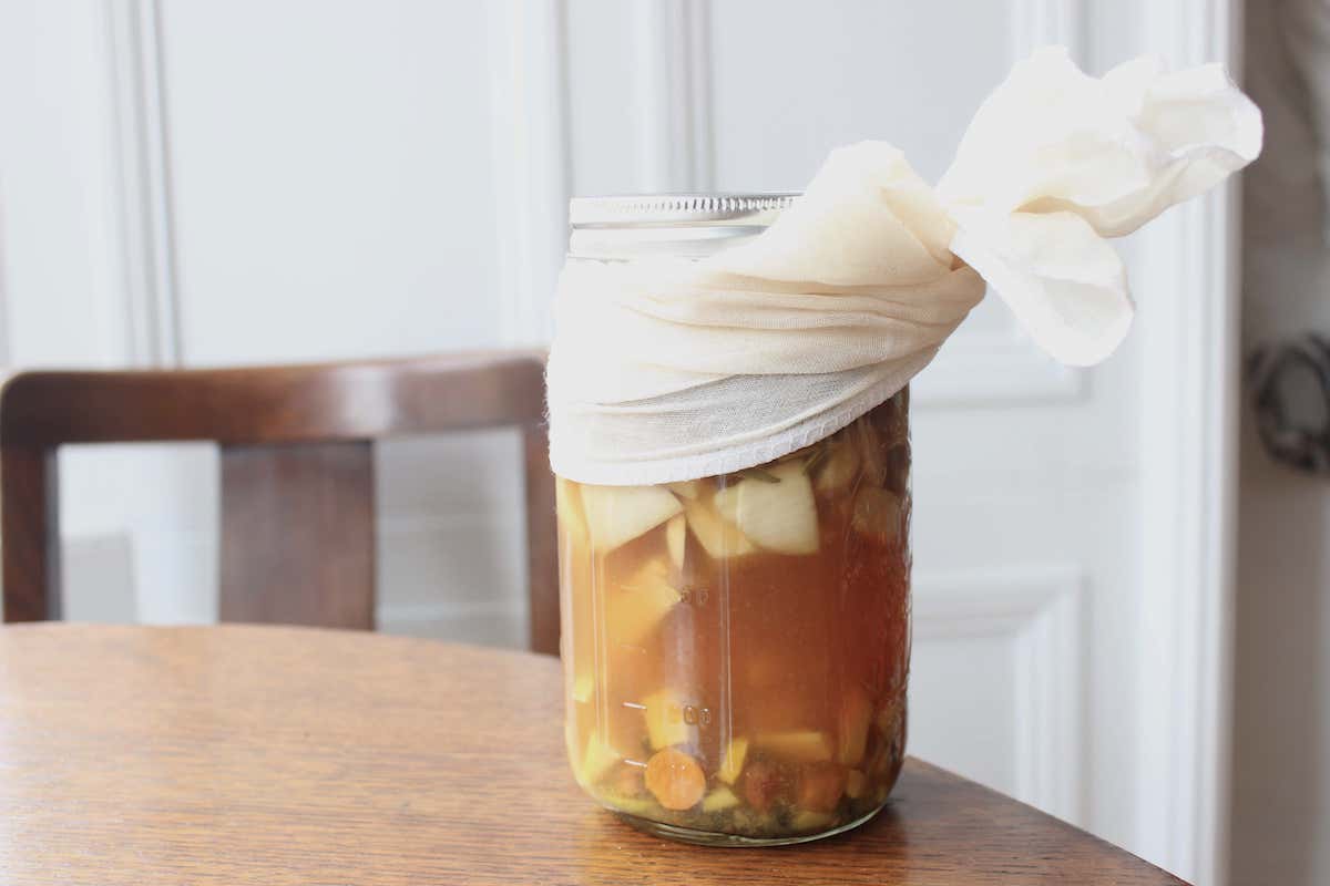 Yellow fire cider in a glass jar with white cheesecloth on top sitting on a wooden table.
