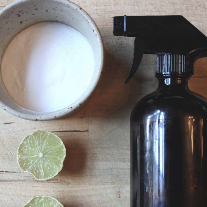 An amber spray bottle on a wooden cutting board next to a small pottery bowl of baking soda and limes.