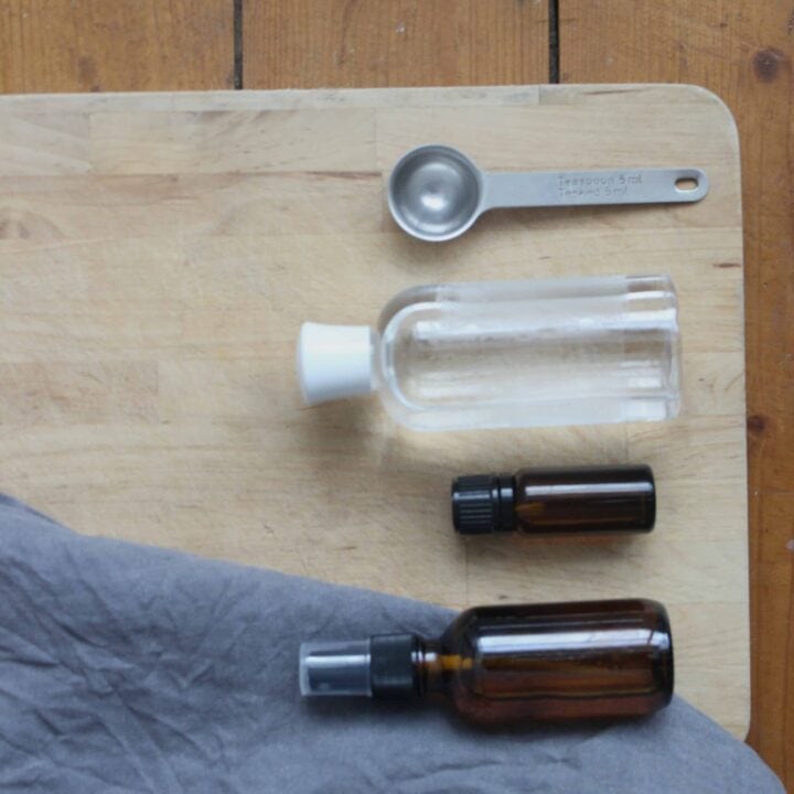 A teaspoon, clear bottle of witch hazel, eucalyptus essential oil, and an amber spray bottle of diy eucalyptus linen spray on a wooden cutting board. There is a wooden floor in the background.