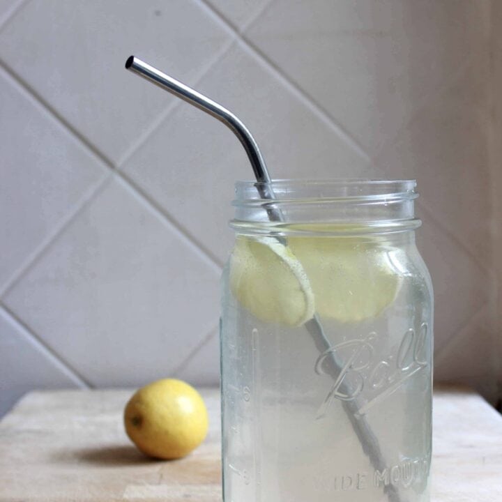 A glass mason jar filled with labor aid electrolyte drink and lemon slices with a stainless steel straw. A lemon is in the background.