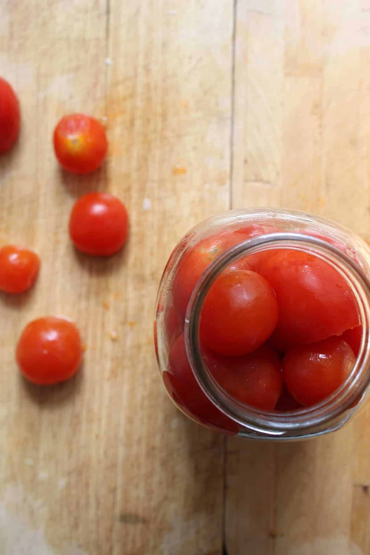 A glass jar of tomatoes on a wooden cutting board with tomatoes beside it.