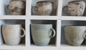 Six handmade Mud Station mugs on white wooden shelves.