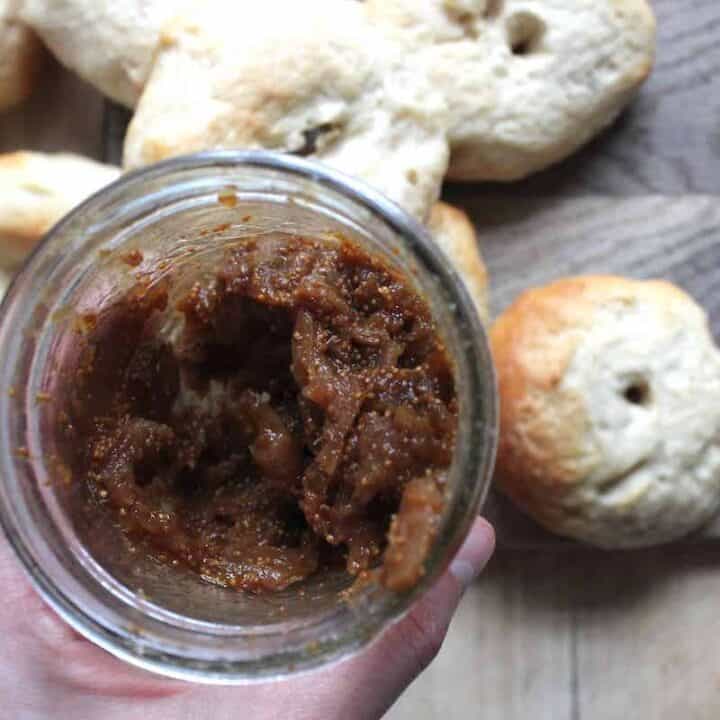 An overhead shot of a hand holding a glass jar of homemade fig jam. There are bagels in the background on a wooden cutting board.
