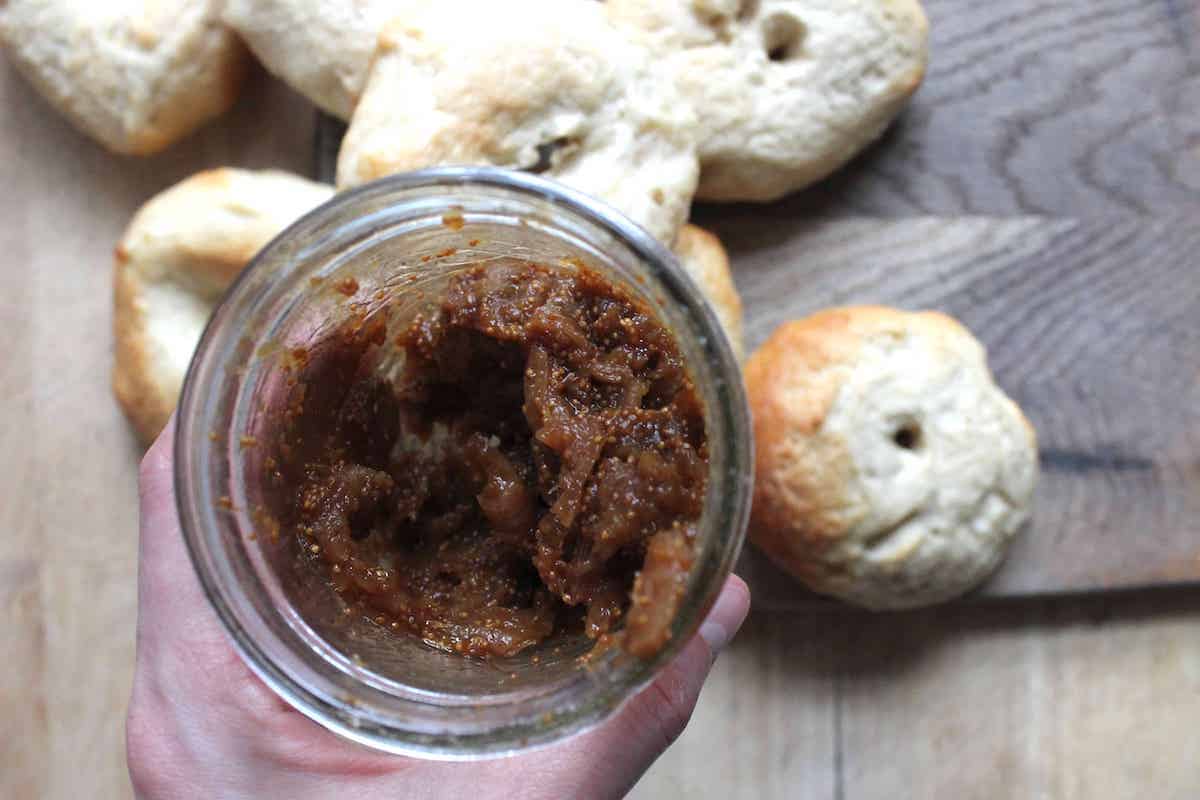 An overhead shot of a hand holding a glass jar of homemade fig jam. There are bagels in the background on a wooden cutting board.