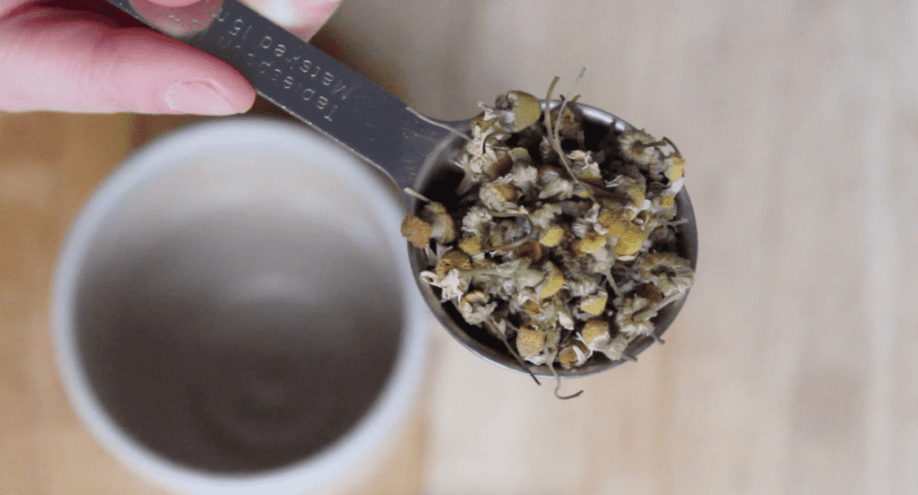 An overhead shot of chamomile flowers in a tablespoon. There is a mug in the background.