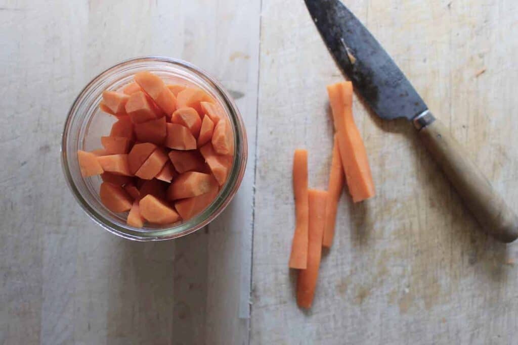 A glass jar with chopped carrots. There is a knife lying next to the jar on a wooden cutting board.