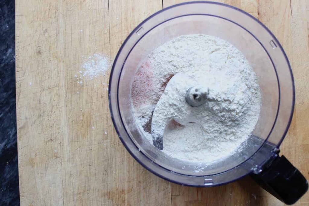 An overhead shot of a food processor on a wooden countertop. There is flour and salt in the food processor.