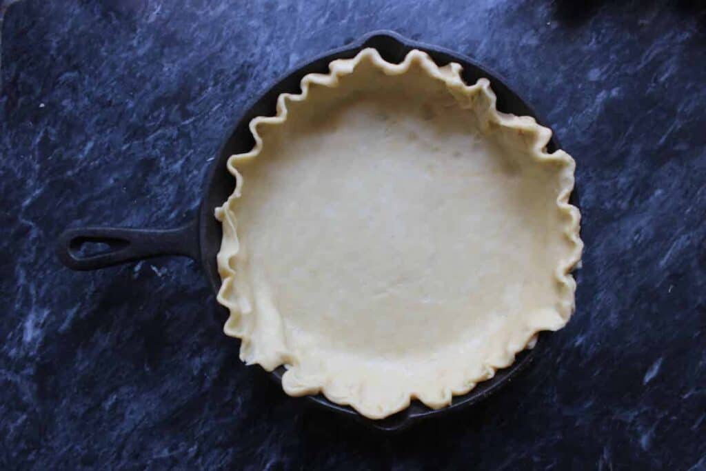 An overhead shot of a cast iron skillet with a homemade pie crust in it. It is sitting on a dark countertop.