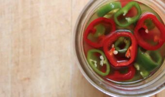 An overhead shot of sliced jalapenos in a saltwater brine in a glass jar on a wooden cutting board.