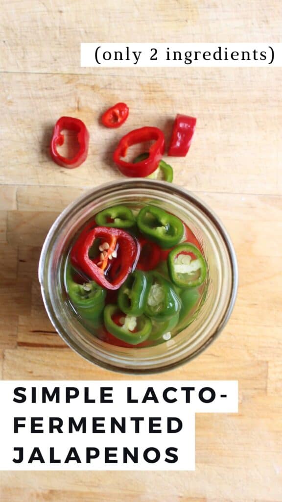 An overhead shot of sliced jalapenos in a saltwater brine in a glass jar on a wooden cutting board. There are sliced jalapenos beside the jar