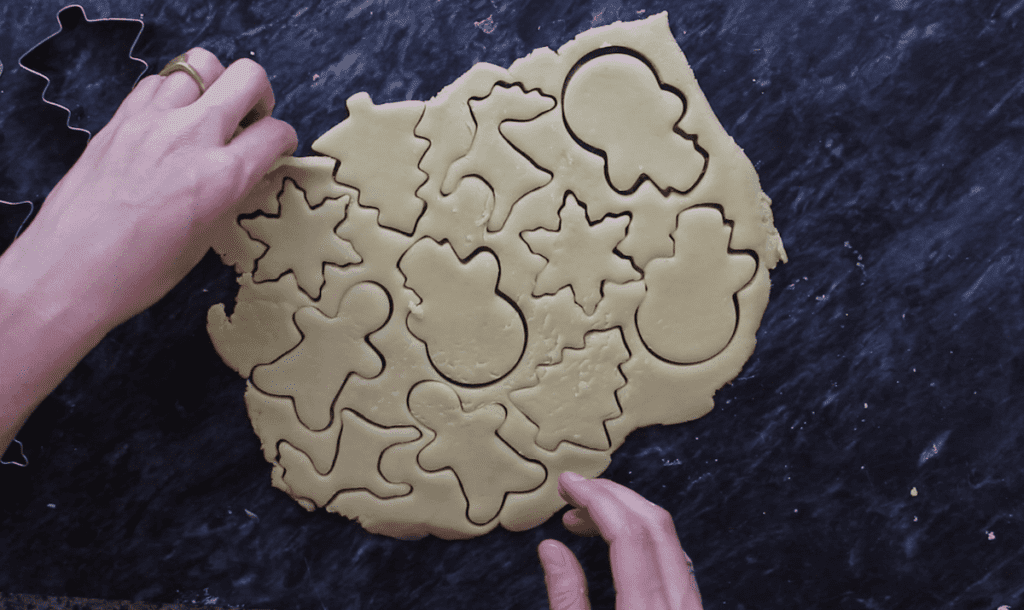 An overhead shot of two hands cutting out sugar cookies from dough on a dark background.