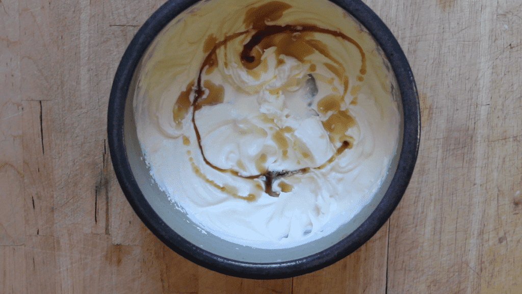 An overhead shot of whipped cream with maple syrup and vanilla that needs to be mixed. The mixure is in a blue bowl on a wooden countertop.
