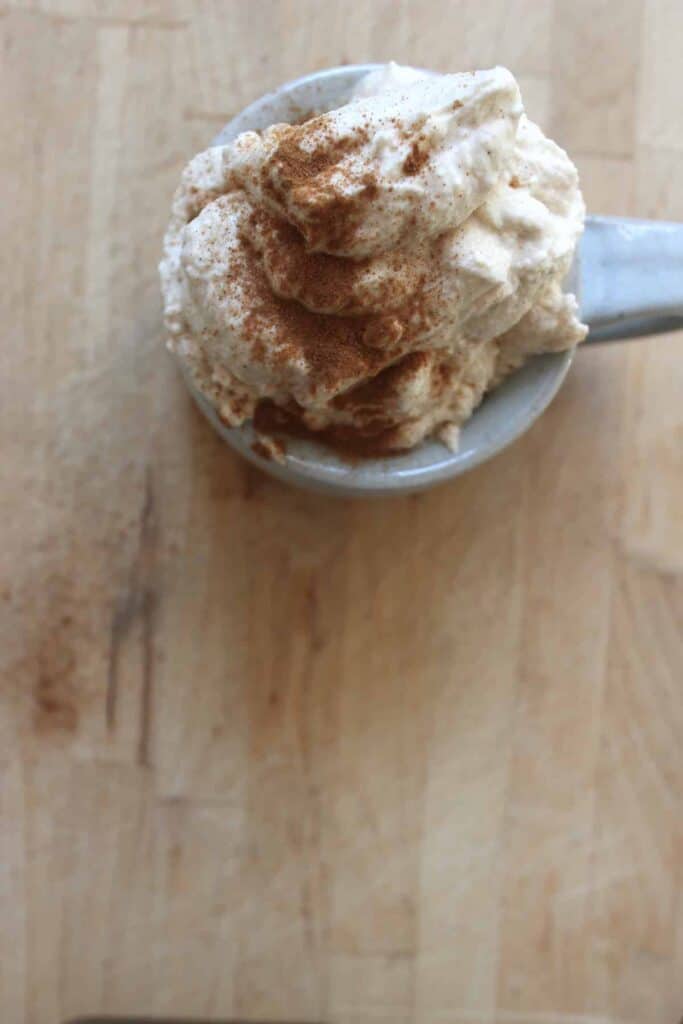 An overhead shot of a grey mug with cinnamon whipped cream in it. There is cinnamon sprinkled on top and the mug is sitting on a wooden countertop.