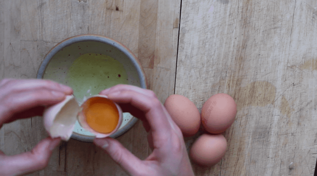 An overhead shot of two hands cracking an egg into a pottery bowl on a light countertop.