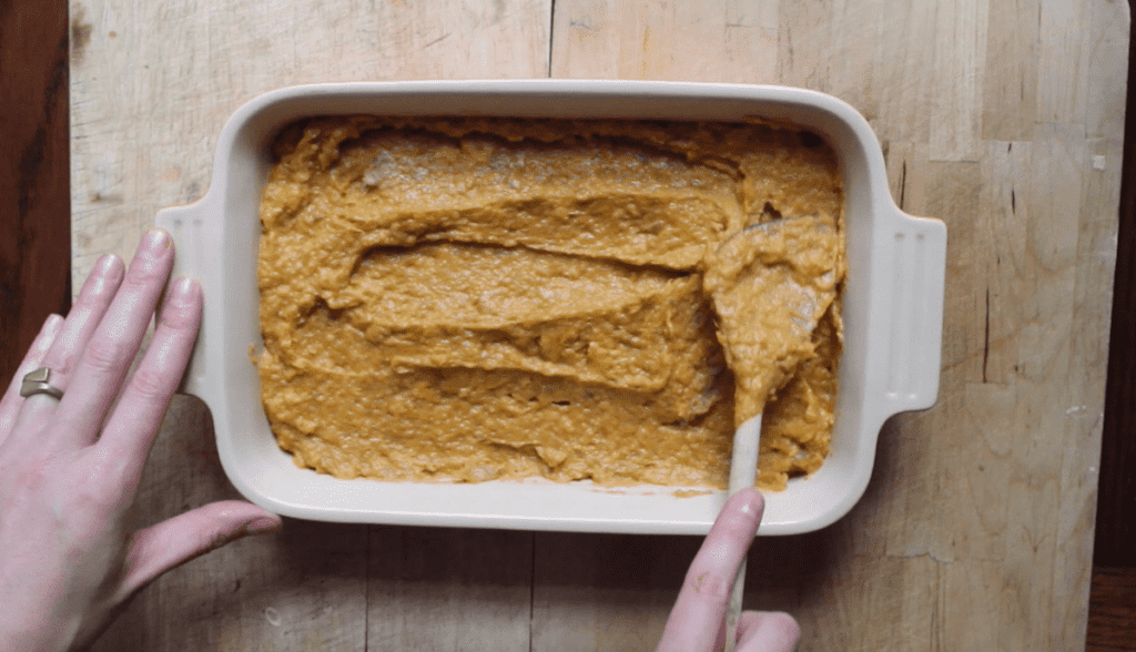An overhead shot of mashed sweet potatoes with maple syrup, eggs, salt, and vanilla in a cream baking dish. A person is spreading it flat with a wooden spoon.