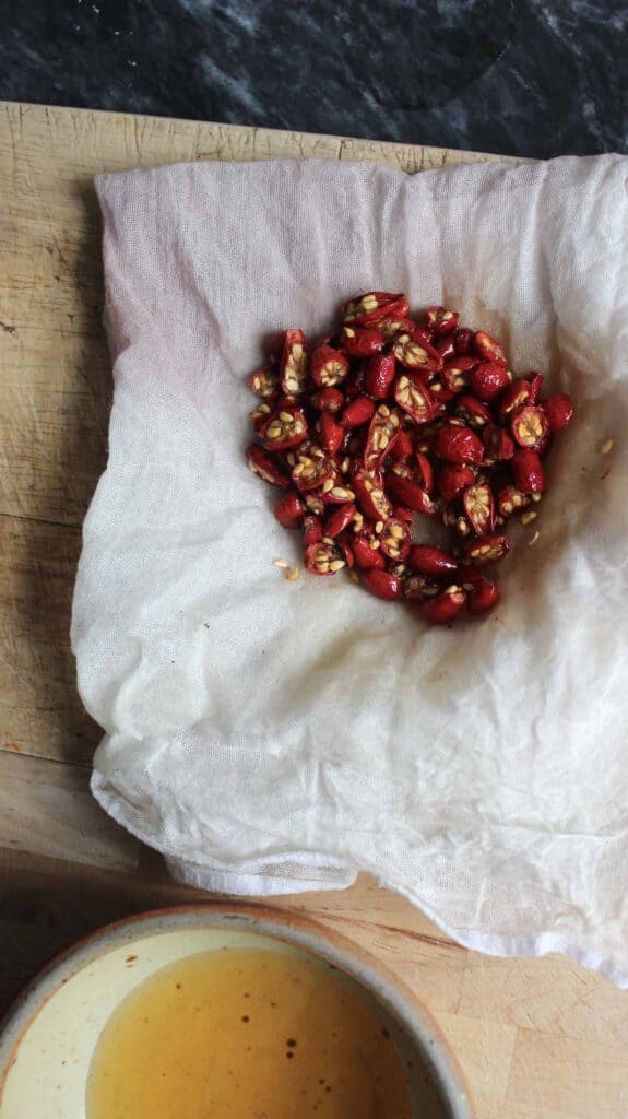 An overhead shot of dried red rosehips on white cheesecloth next to a mall bowl of rosehip oil. They are on a light countertop.