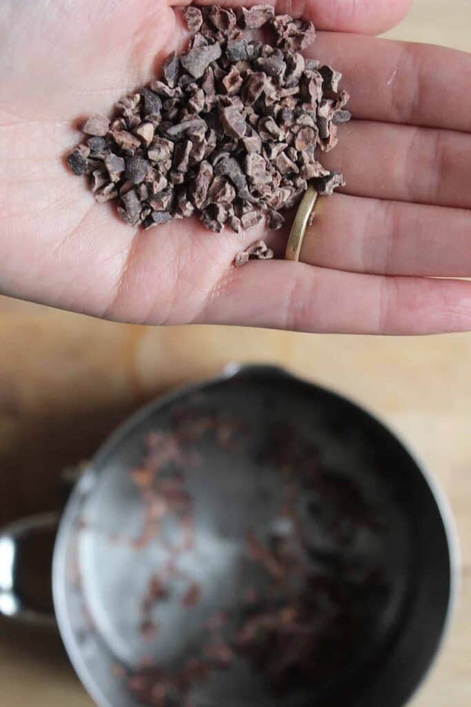 An overhead shot of a hand holding raw cacao nibs. There is a silver saucepan in the background.