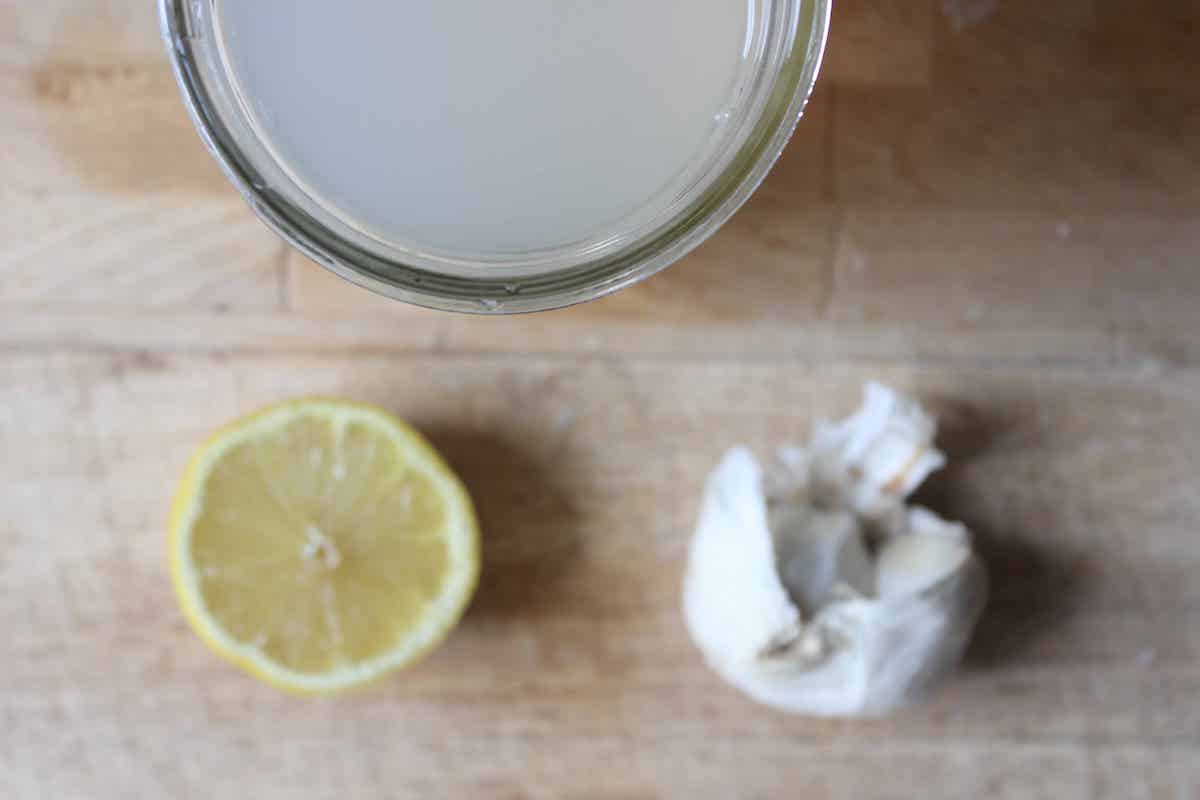 An overhead shot of half a lemon, a bulb of garlic, and a glass jar of garlic tea on a wooden cutting board.