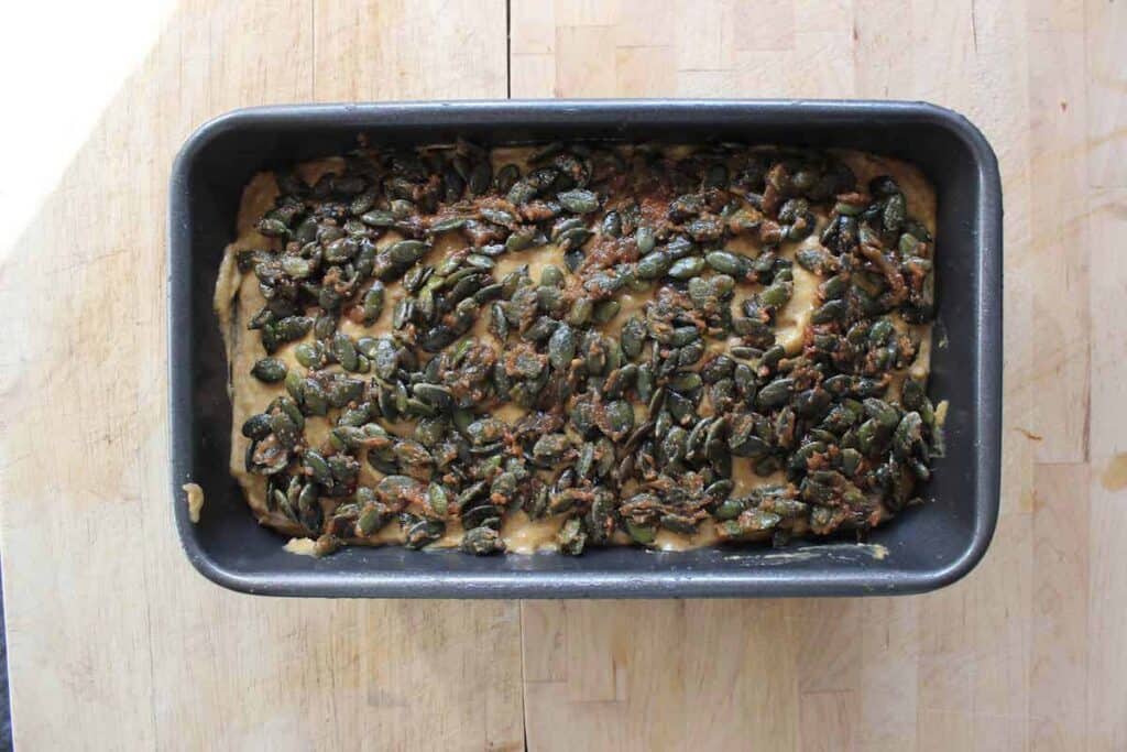 An overhead shot of unbaked sourdough pumpkin bread batter in a loaf tin with sugary pumpkin seeds on top.