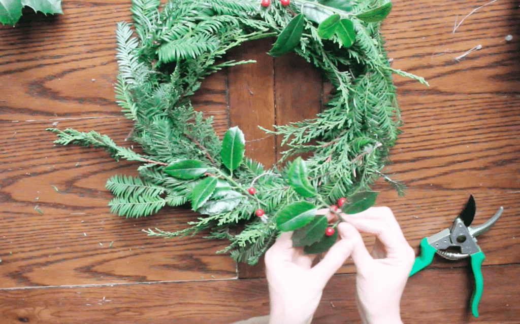 An overhead shot of a person hot gluing foraged holly to a wreath form.