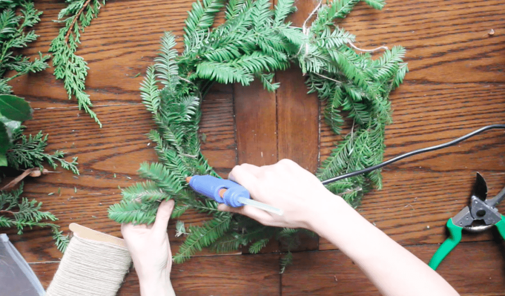 An overhead shot of a person hot gluing foraged greenery to a wreath form.