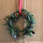A foraged wreath with greenery, red berries, dried oranges, and red ribbon hanging on a wooden door.