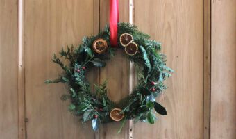 A foraged wreath with greenery, red berries, dried oranges, and red ribbon hanging on a wooden door.