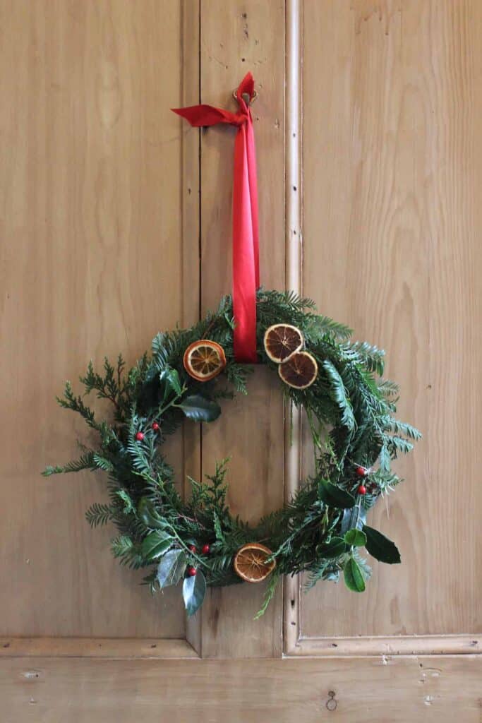 A foraged wreath with greenery, red berries, dried oranges, and red ribbon hanging on a wooden door.
