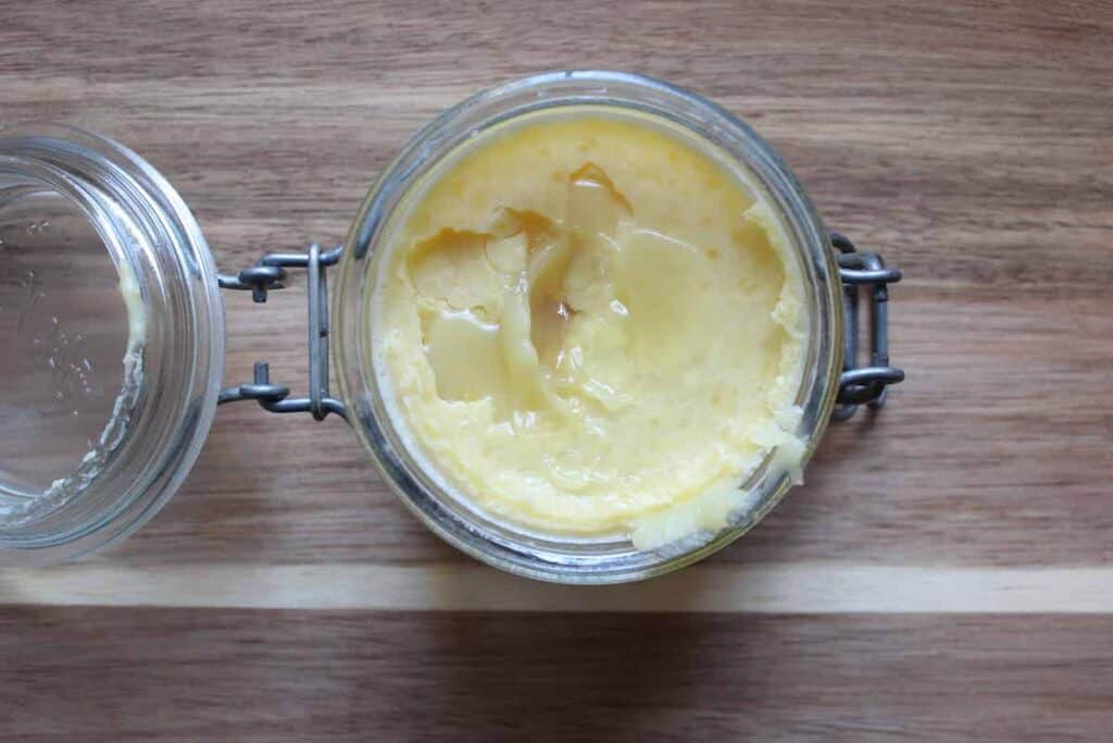 An overhead shot of ﻿﻿beeswax, coconut oil, shea butter, and cocoa butter combined into a body butter in a glass jar on a light countertop.