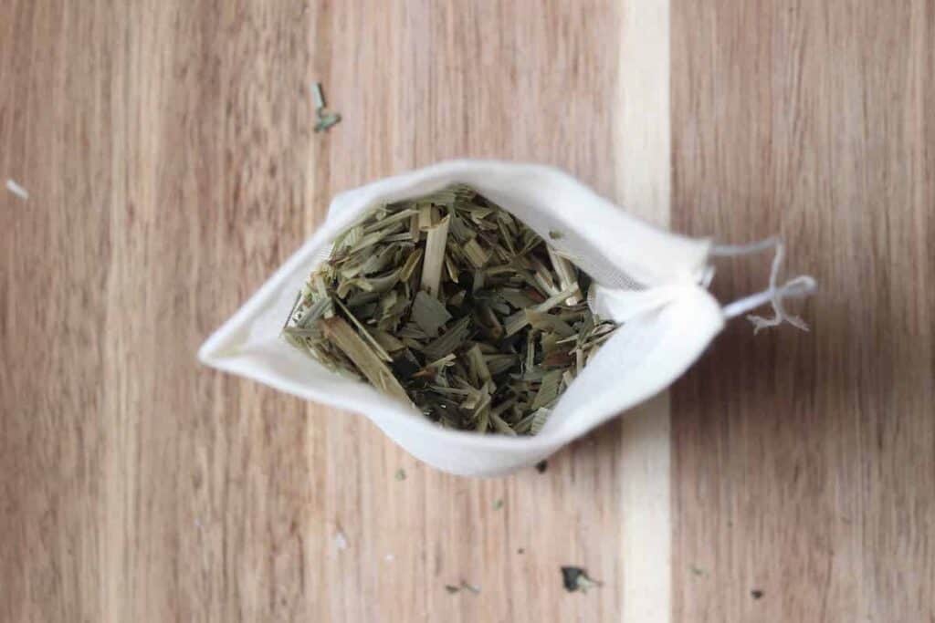 An overhead shot of dried oat straw and nettles in a small muslin bag on a light countertop.