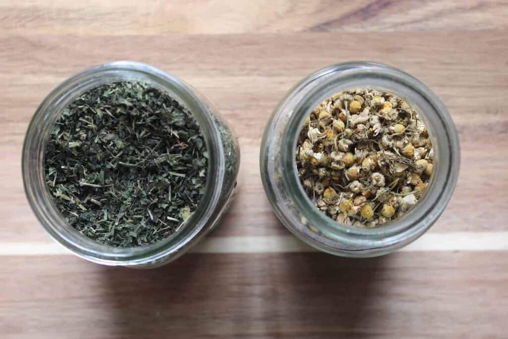 An overhead shot of two glass jars with dried green herbs and dried chamomile on a light countertop.