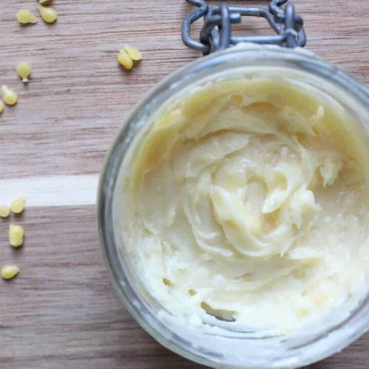 An overhead shot of ﻿﻿beeswax, coconut oil, shea butter, and cocoa butter whipped into a body butter in a glass jar on a light countertop. There are beeswax pellets beside the jar.