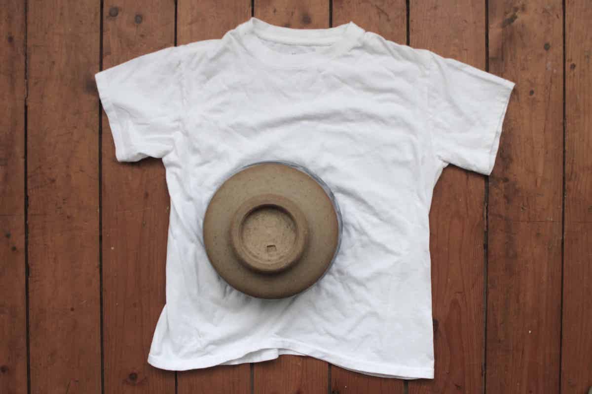 An overhead shot of a white tshirt with a small bowl turned upside down on top. The tshirt is lying on a wooden floor.