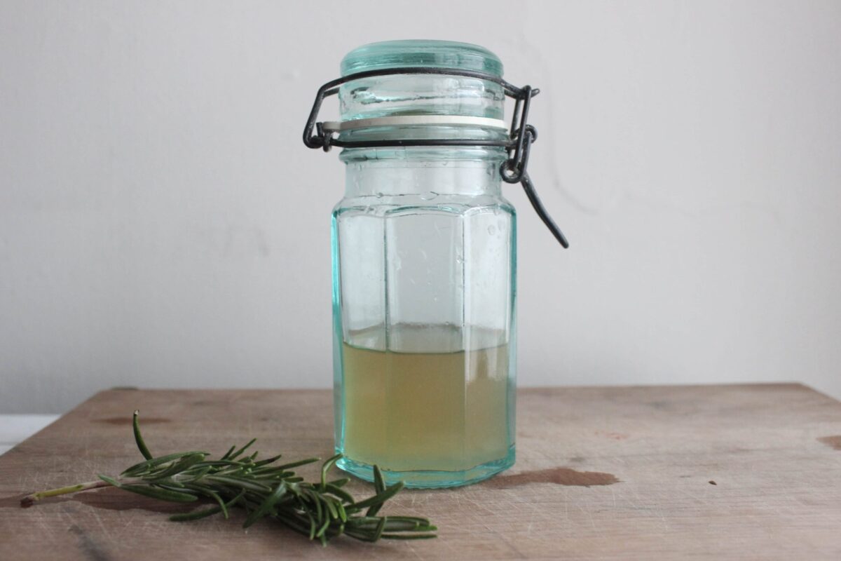 A vintage green and blue jar glass with a few sprigs of fresh rosemary in front of it. There is rosemary tincture in the glass jar.