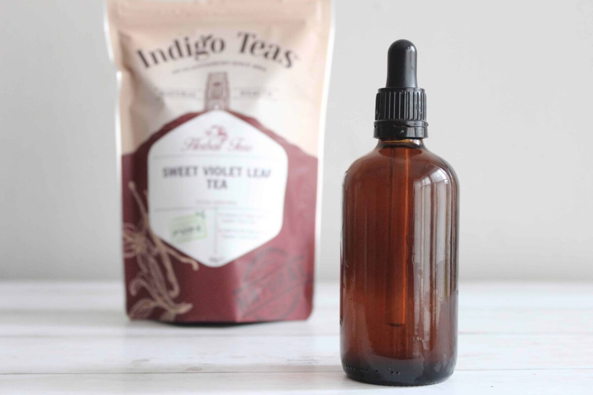 An amber glass bottle with violet tincture inside. There's a bag of violet leaves behind the bottle.