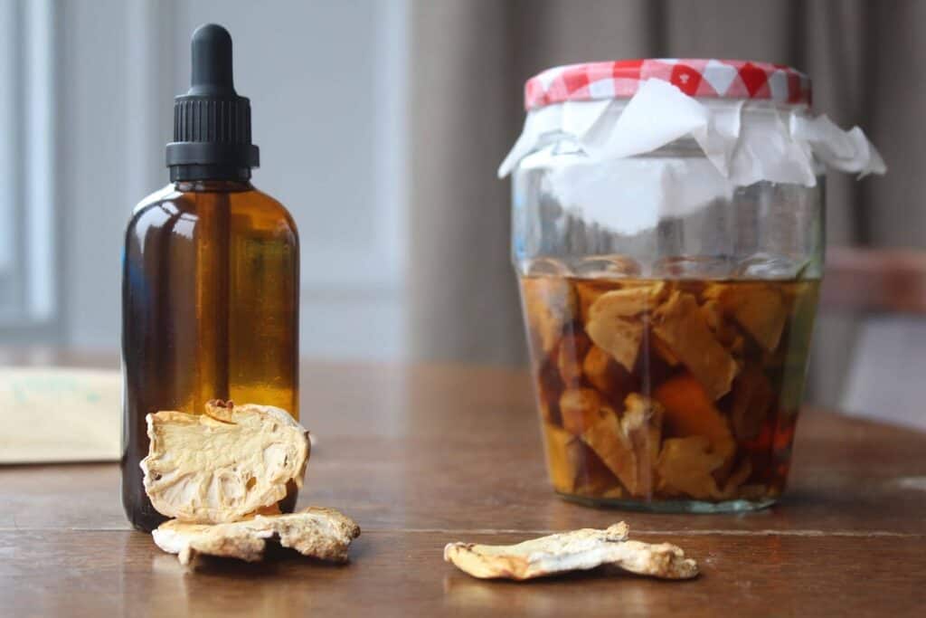 An amber glass dropper bottle with lion's mane tincture inside. There's a glass jar with lion's mane steeping in alcohol beside the tincture bottle. There are white chunks of lion's mane in front of the dropper bottle.