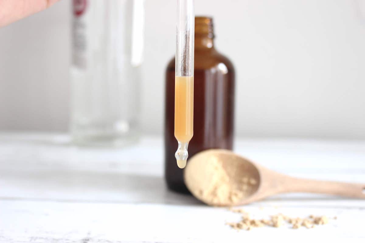 Lion's Mane tincture in a small amber glass bottle. There is a bottle of vodka in the background and a wooden tablespoon with powdered Lion's Mane mushroom in the teaspoon. A dropper with the tincture is being held in front of everything.