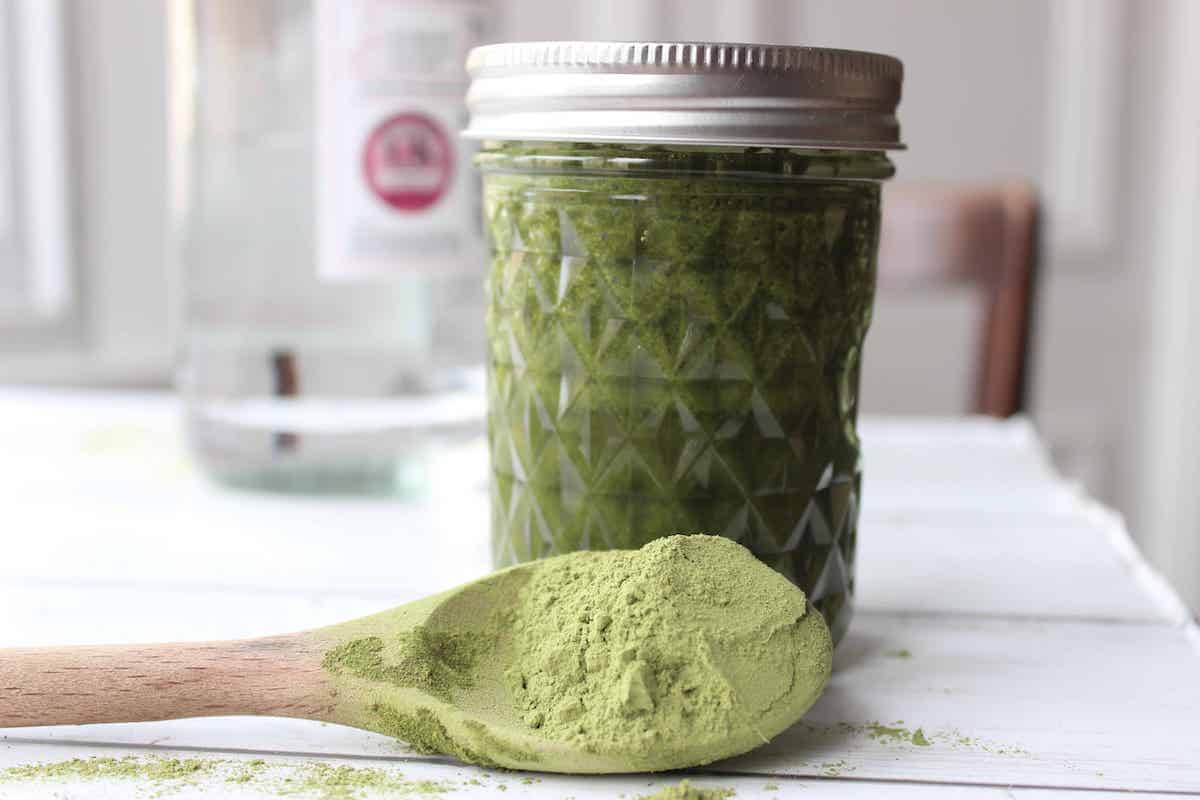 Moringa tincture steeping in a glass jar. There is a wooden spoon with moringa powder in front of the jar.