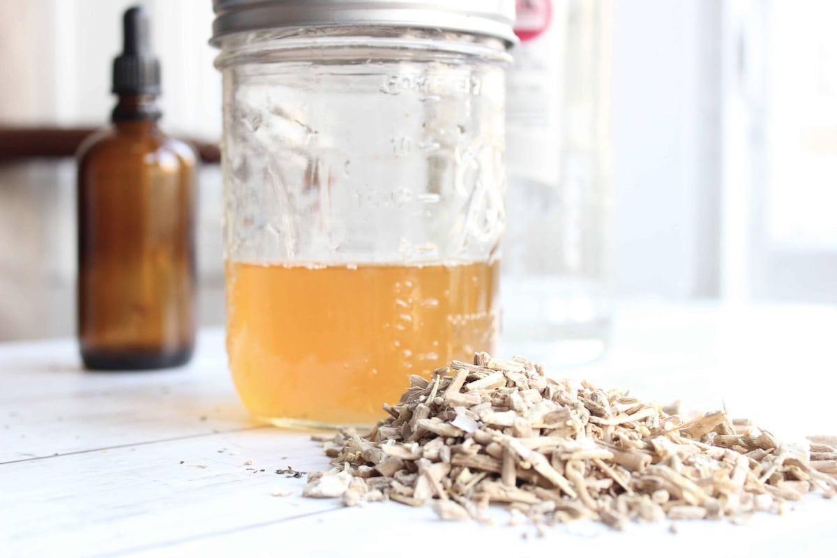 A glass jar with ashwagandha tincture inside. There is dried ashwagandha in front of it and an amber glass tincture bottle in the background.