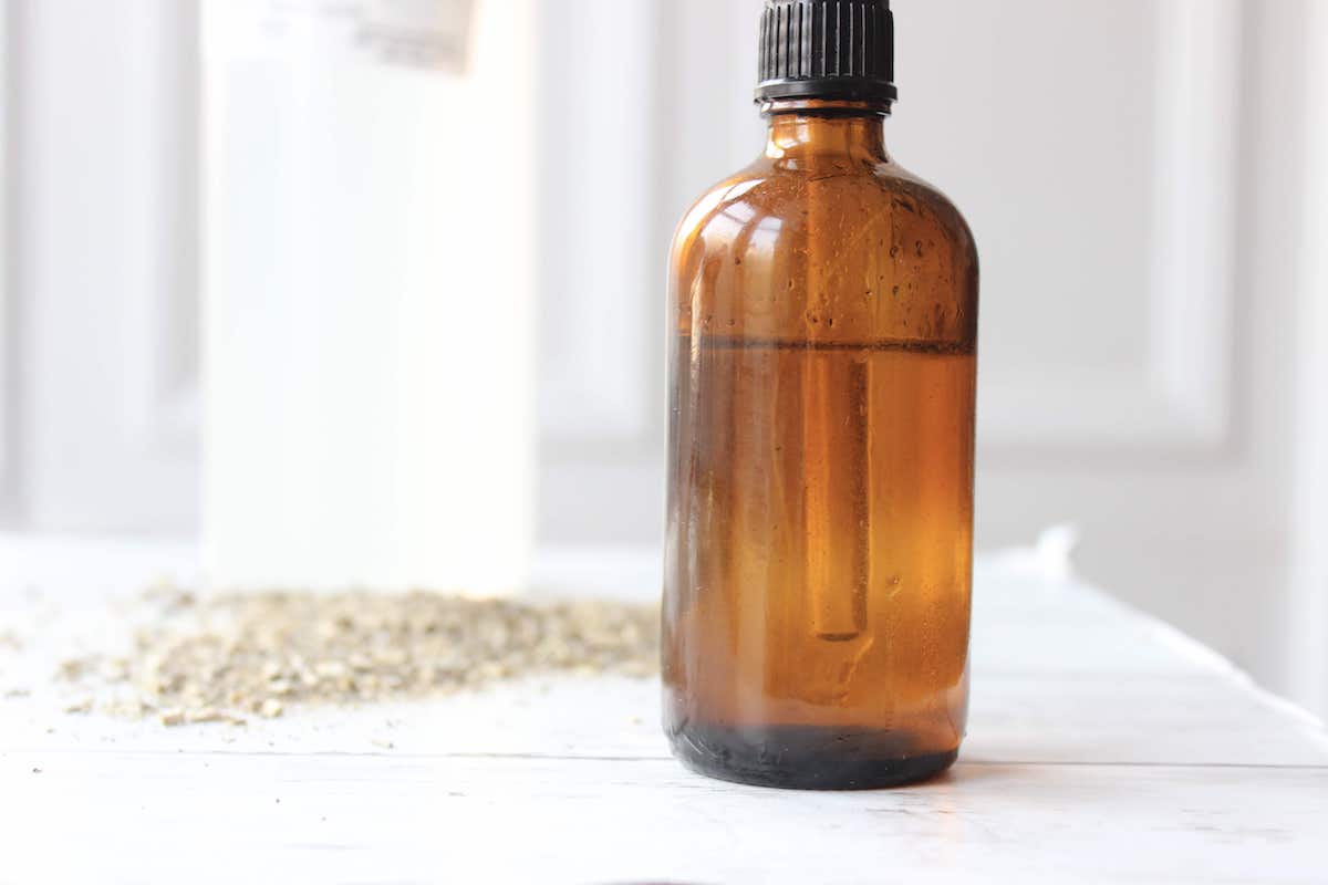 A close up of an amber glass dropper bottle with dried herbs behind it and a bottle of food-grade glycerin.