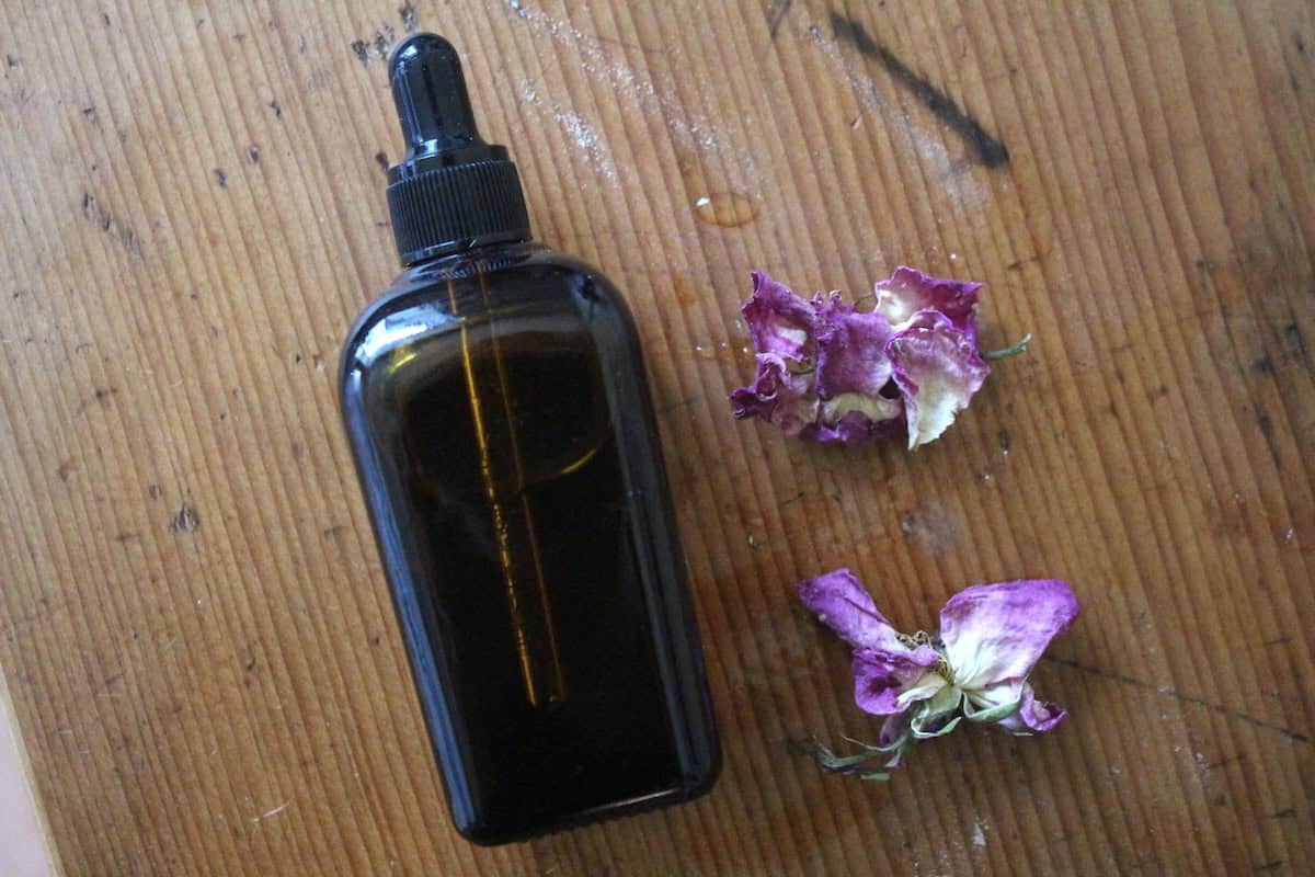 An overhead shot of an amber glass oil dropper bottle with roseoil inside and dried roses next to it.