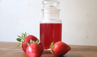 A small glass jar with red strawberry cordial inside and fresh strawberries in front of it.