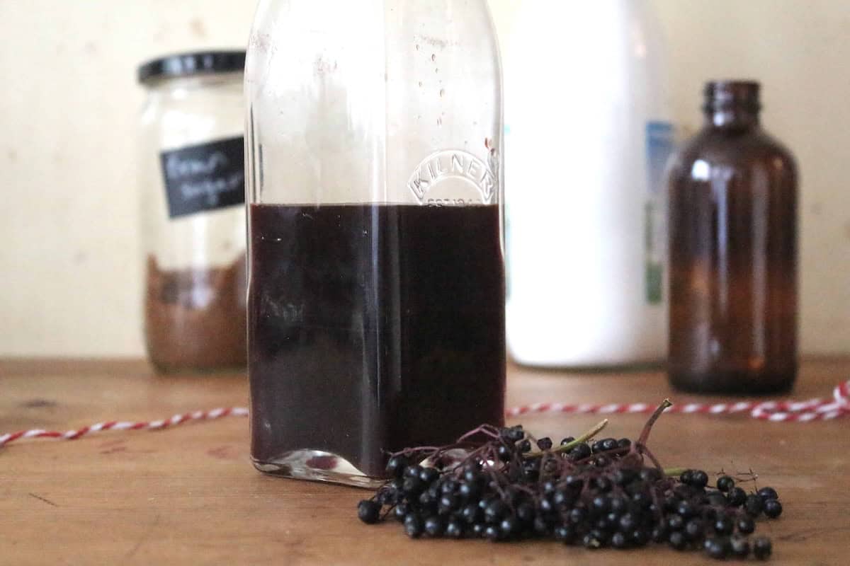 A glass swing top bottle with elderberry cordial inside. There's a small amber glass bottle in the background, next to a large white bottle and a glass jar with brown sugar. There are fresh elderberries in front of the cordial.
