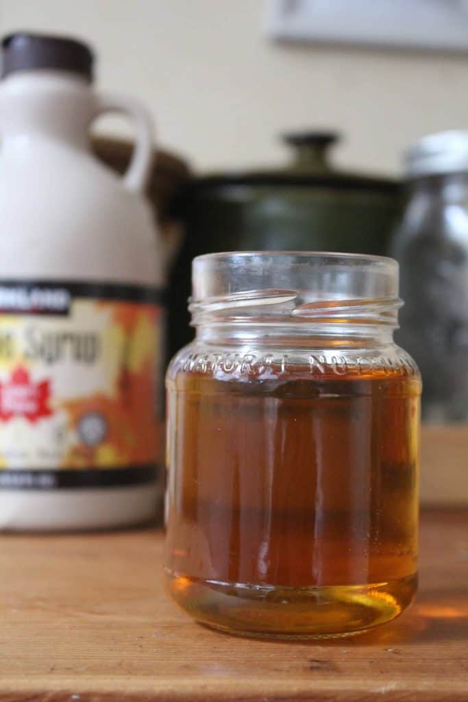 A small glass jar with maple simple syrup inside. There's a bottle of maple syrup in the background.