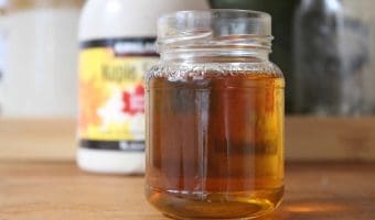 A small glass jar with maple simple syrup inside. There's a bottle of maple syrup in the background.
