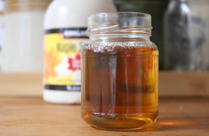 A small glass jar with maple simple syrup inside. There's a bottle of maple syrup in the background.