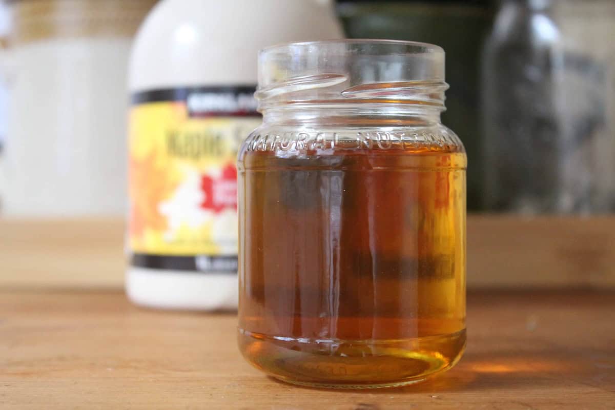 A small glass jar with maple simple syrup inside. There's a bottle of maple syrup in the background.